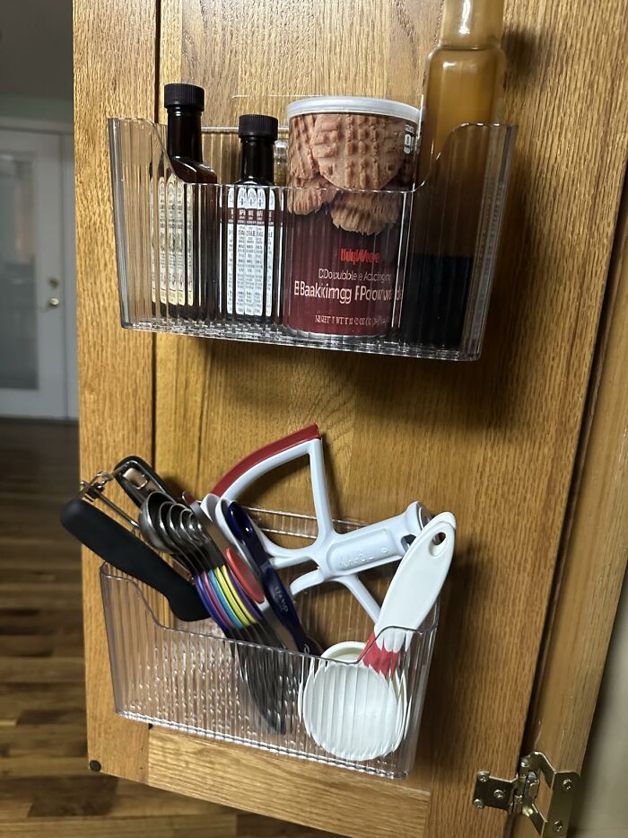 Clear kitchen organizer mounted on a cabinet door storing measuring spoons, peelers, oil bottles, and other kitchen items.