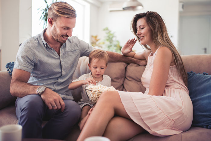 Child-free lady warning sister about too much babysitting while sitting on couch with man and child sharing popcorn.