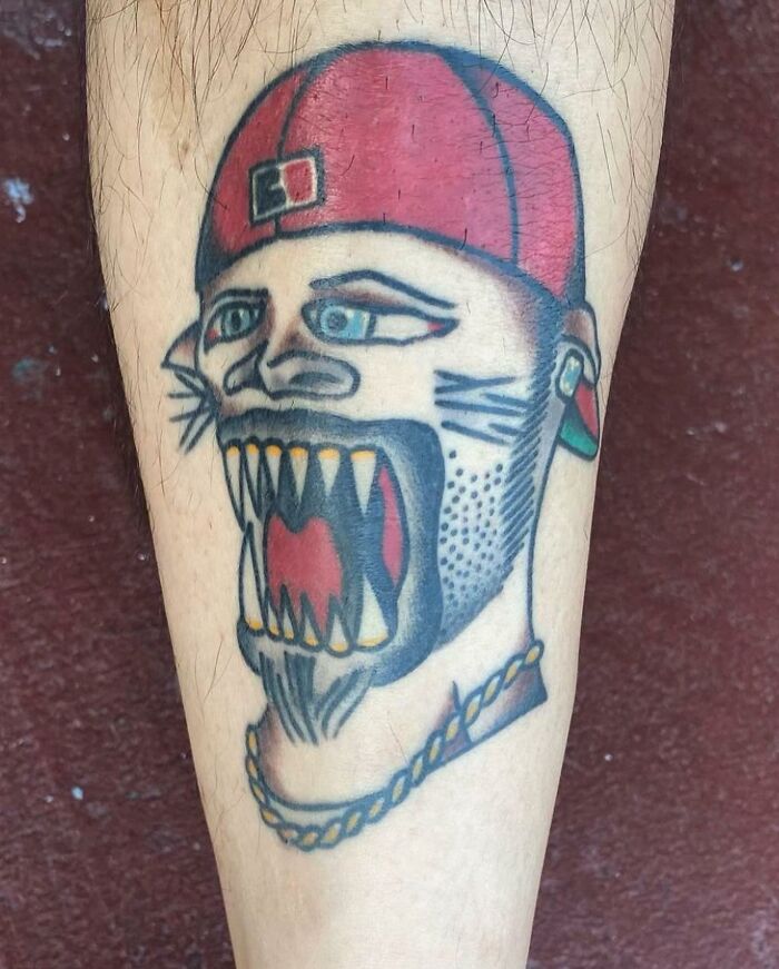 Tattoo of a stylized face with sharp teeth, wearing a red cap and a gold chain, showing bizarre choices people made tattoos.