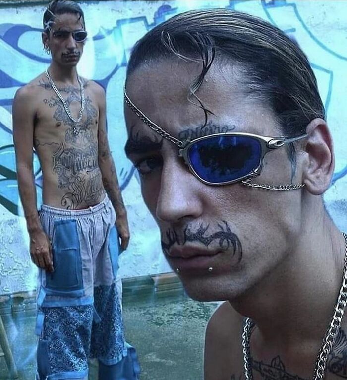 Man with bizarre face and chest tattoos wearing sunglasses with chain and patchwork pants in urban setting.
