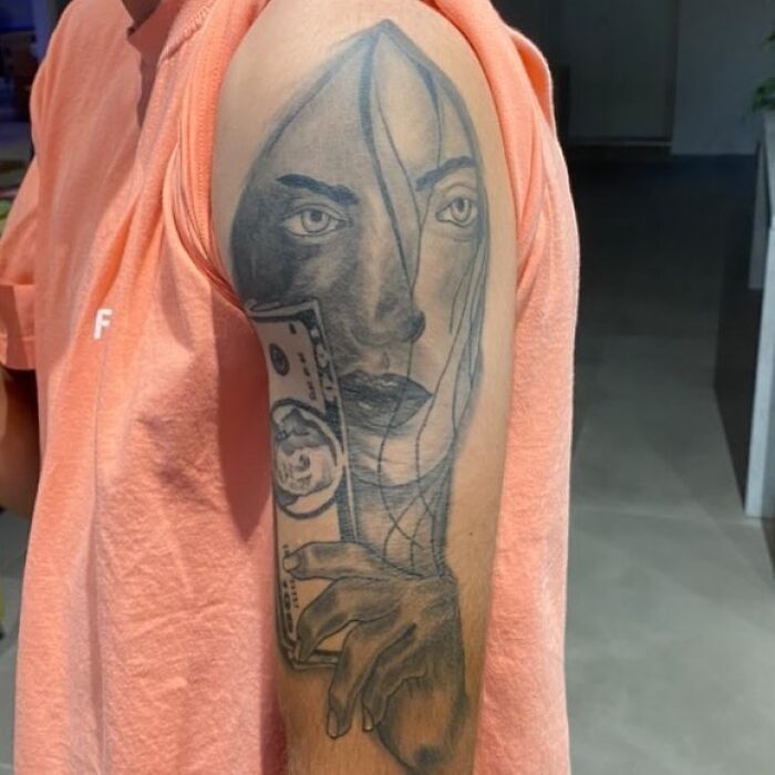 Tattoo of a surreal face holding money on upper arm, showcasing bizarre choices people made while getting tattoos.