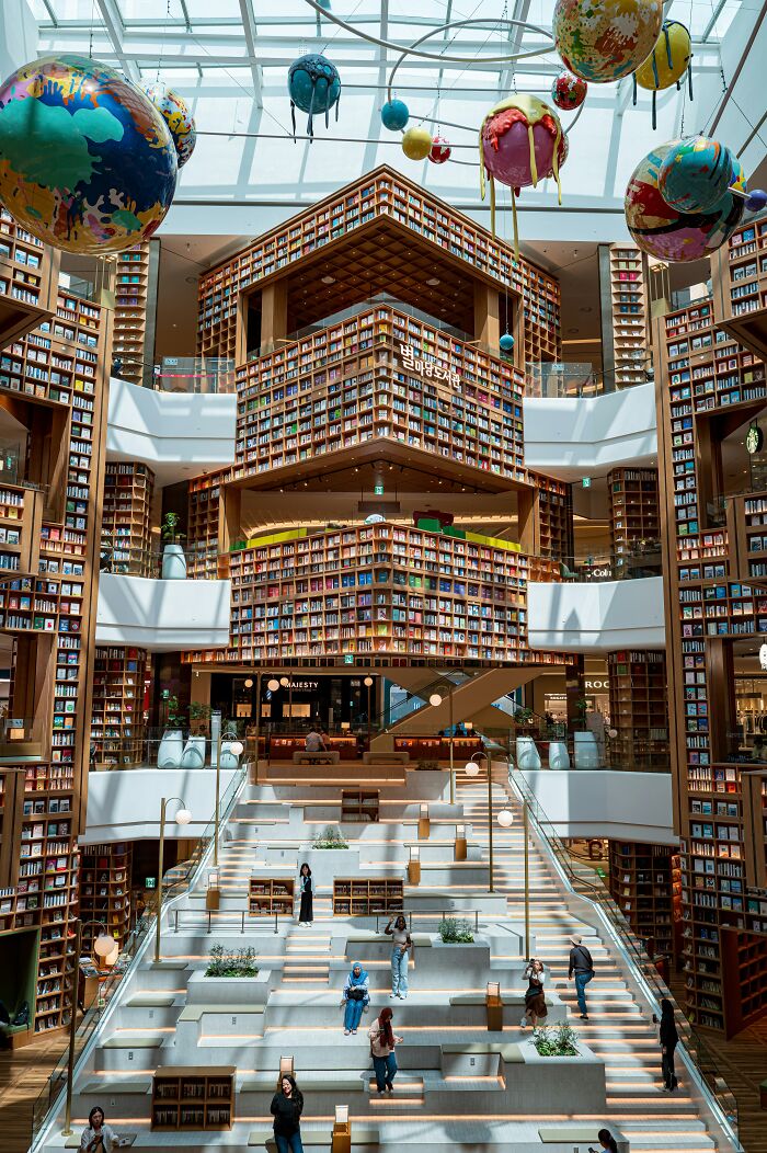 Stunning bookstore interior with towering shelves, large colorful hanging globes, and visitors on stepped seating area under glass ceiling.