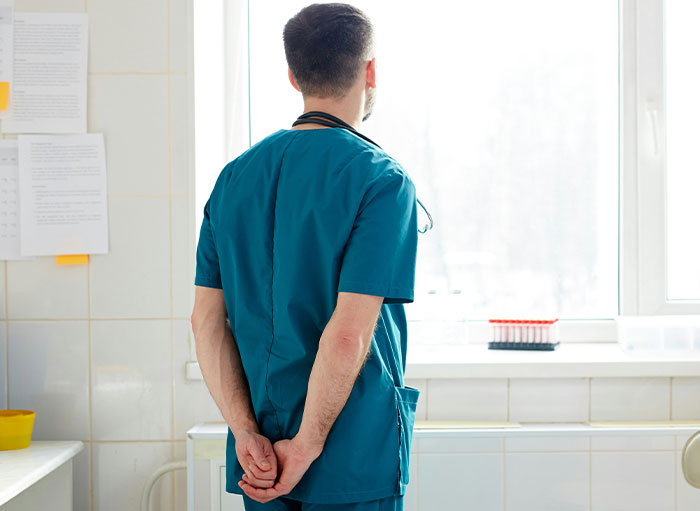 Surgeon in blue scrubs standing by a bright window with arms behind back in a clinical medical setting.