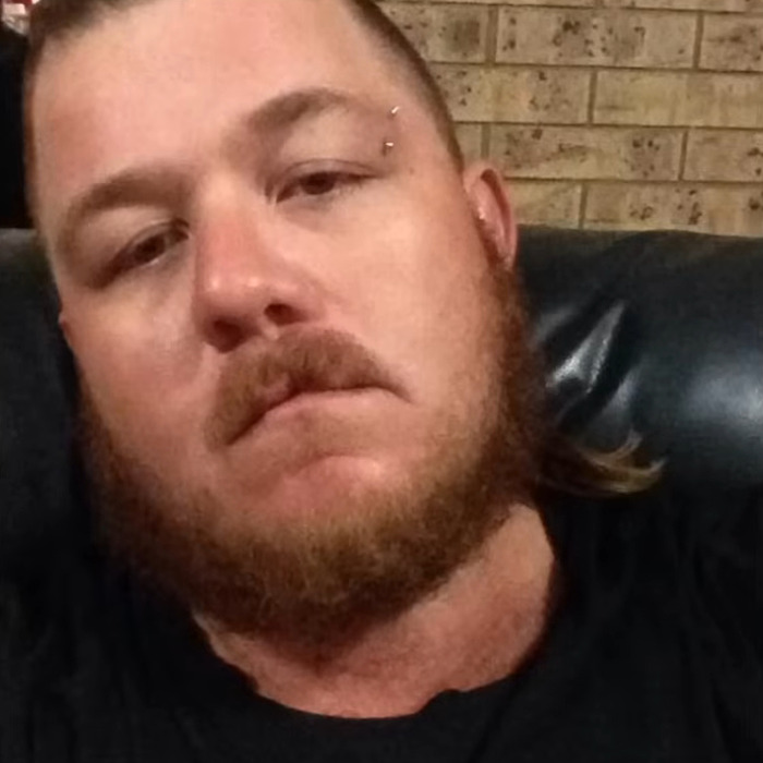 Bearded man with eyebrow piercing sitting on a black leather couch against a brick wall background.