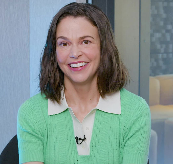 Sutton Foster smiling during an interview, wearing a green sweater and white collared shirt indoors. Sutton Foster smiling during an interview, wearing a green sweater and white collared shirt indoors.