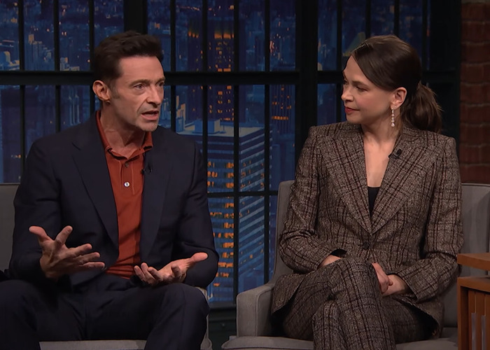 Hugh Jackman speaking animatedly beside a woman in a brown suit during a late-night talk show interview.