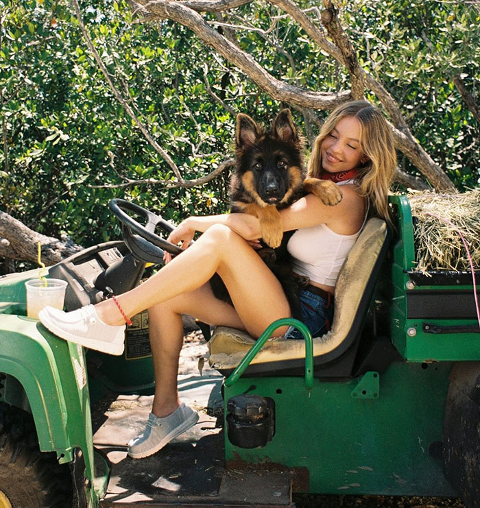 Sydney Sweeney sitting on a green vehicle outdoors, holding a German Shepherd puppy during a daring photoshoot.
