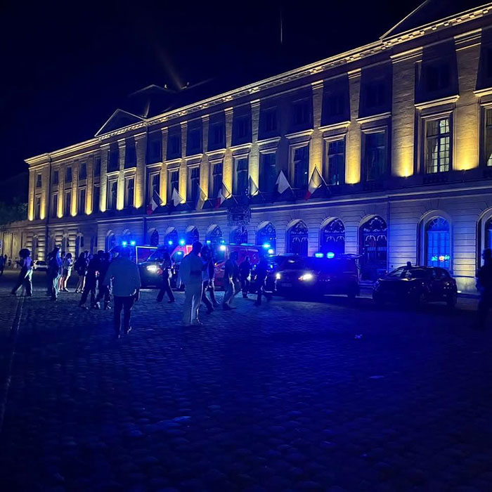 Crowd and emergency vehicles with flashing lights gathered at night outside a large lit building after syringe incidents at music festival.
