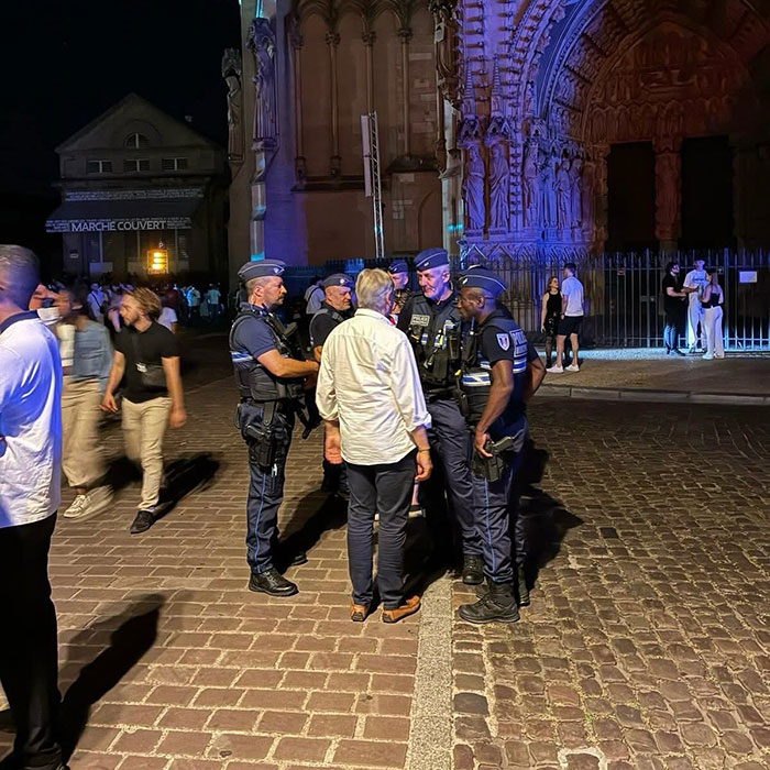 Police officers speaking with a man at night near a historic building amid reports of syringe attacks and violence against women.