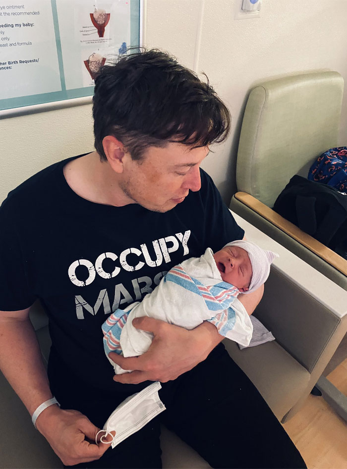 Elon Musk holding newborn baby wrapped in hospital blanket, symbolizing chaotic musk family saga and Tau Techno Mechanicus theme.