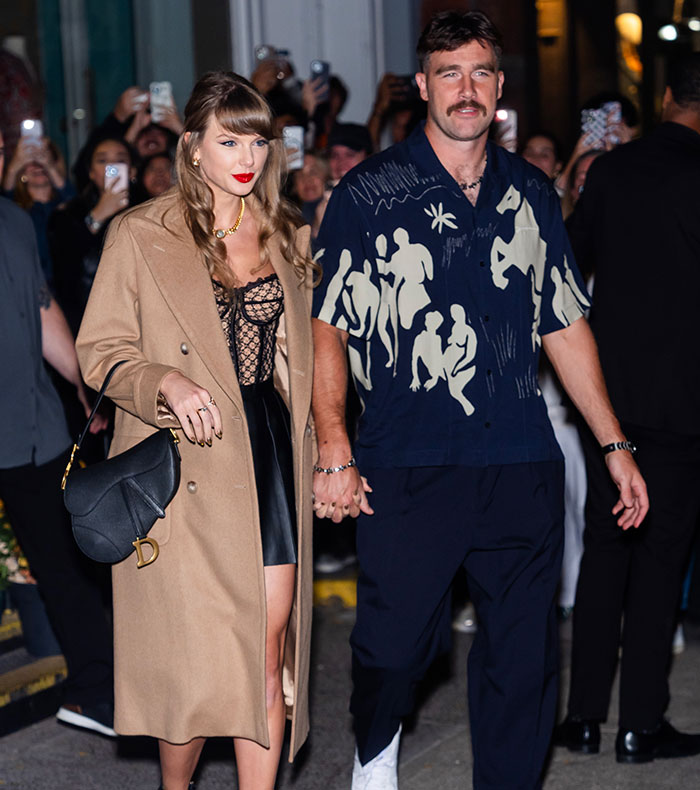 Taylor Swift walking with companion at night, surrounded by fans and photographers with phones raised.