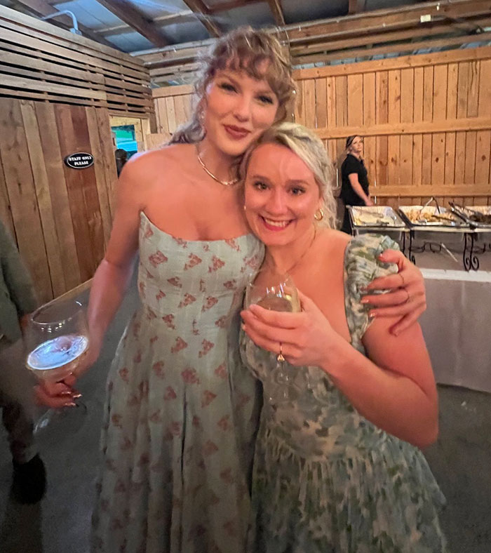 Taylor Swift smiling with a friend, both in floral dresses, at a casual indoor gathering with drinks and food in the background.