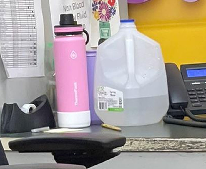 Cluttered office desk with water jug, pink water bottle, tape dispenser, and phone in a toxic work environment photo.