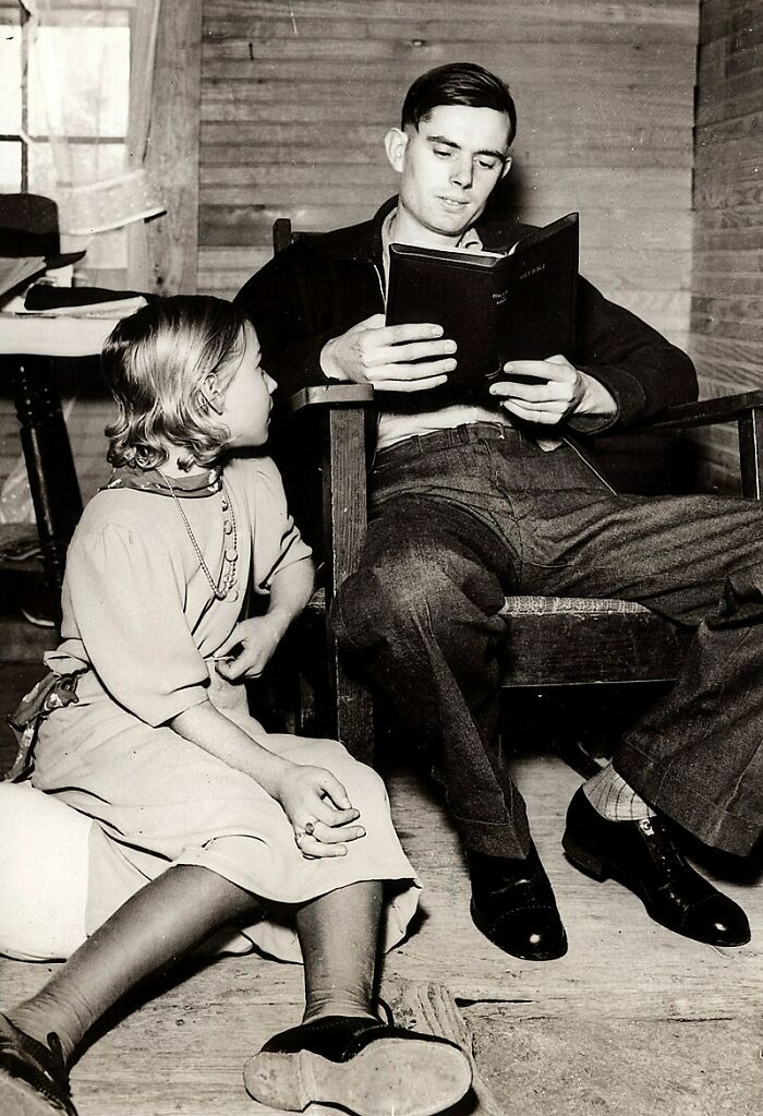 Black and white photo of a young man reading to a girl in a rustic room, an unsettling vintage moment captured from the past.