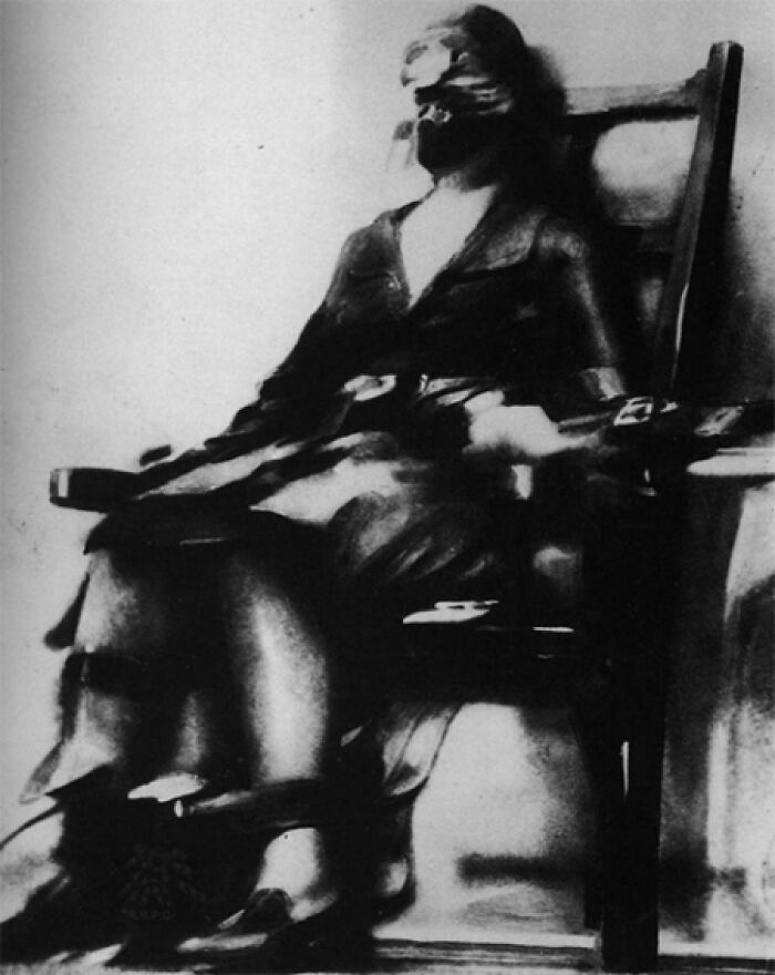Black and white unsettling photo of a person strapped to a chair with an obscured face, creepy vintage image from the past.