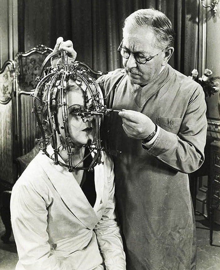 Black and white photo of a man adjusting a creepy metal headgear on a woman in a vintage unsettling medical setting.
