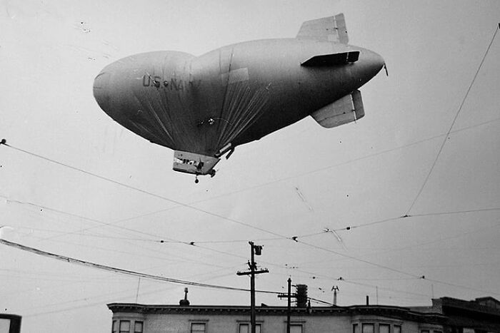 Black and white vintage photo of a US Navy airship flying over buildings with power lines, creepy and unsettling past image.