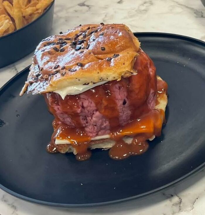 Raw hamburger patty with sauce and melted cheese in a bun on a black plate, one of the disgusting food pics.