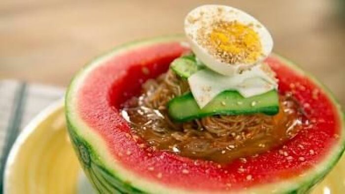 Watermelon bowl filled with noodles, topped with cucumber slices and a halved boiled egg in a disgusting food pic.
