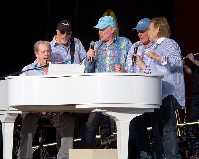 The Beach Boys performing live on stage with a white piano, highlighting legendary co-founder and musical genius.
