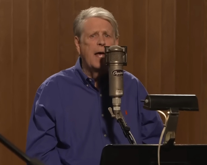 Co-founder of the Beach Boys performing in a studio, wearing a blue shirt and singing into a vintage microphone.