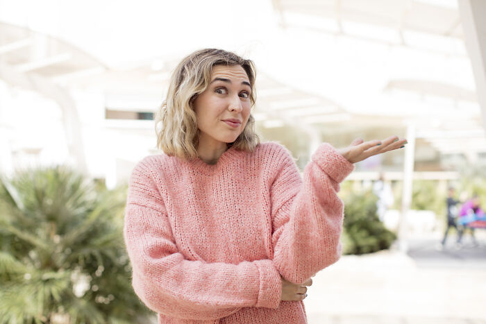 Young woman in a pink sweater shrugging with a curious expression, symbolizing things learned embarrassingly late insights.
