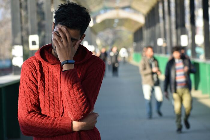 Young man in a red sweater covering his face, feeling embarrassed while standing on a busy outdoor walkway, things learned late