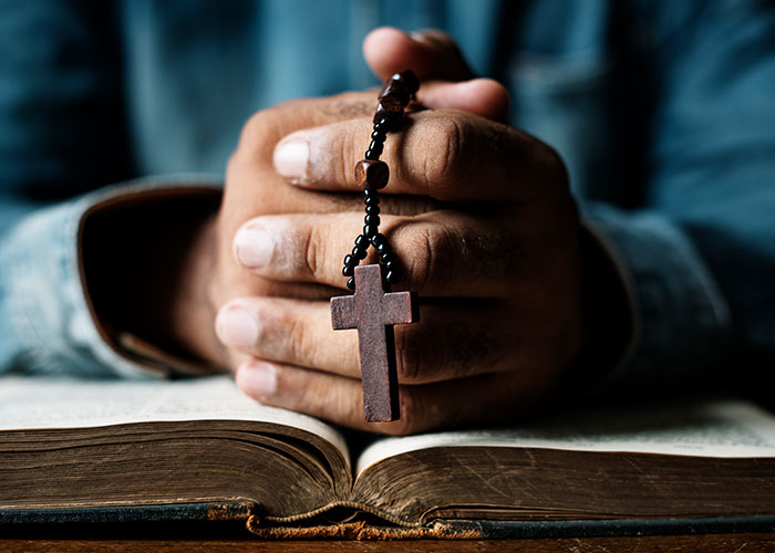 Hands holding a wooden cross necklace over an open book, reflecting on modern behaviors they can’t accept.