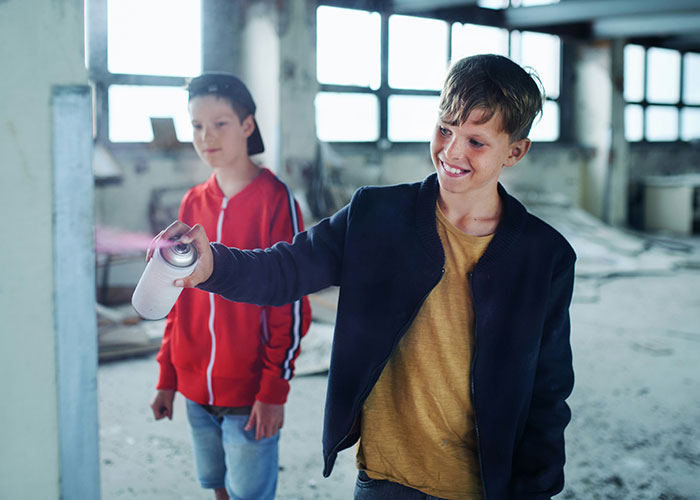 Two boys in an abandoned building, one spraying paint while reflecting on modern behaviors they can’t accept.
