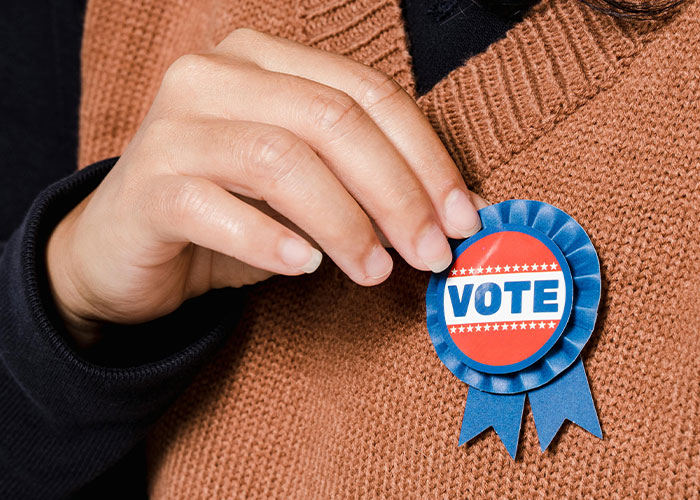 Close-up of a person pinning a vote ribbon on a brown sweater, highlighting modern behaviors and generational perspectives.