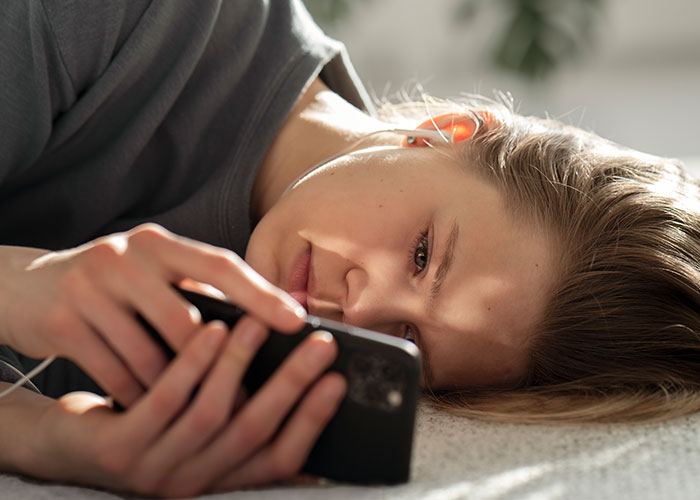 Young woman lying down scrolling on smartphone, illustrating modern behaviors many find hard to accept from how they grew up.
