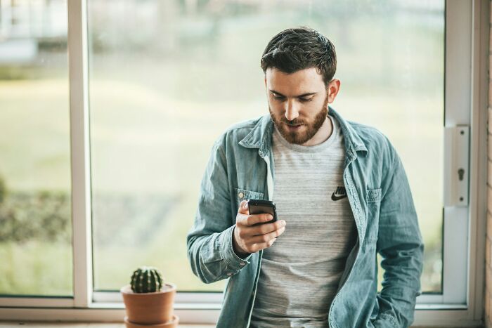 Young man standing by window, focused on his phone, reflecting on personal mysteries and human nature insights.