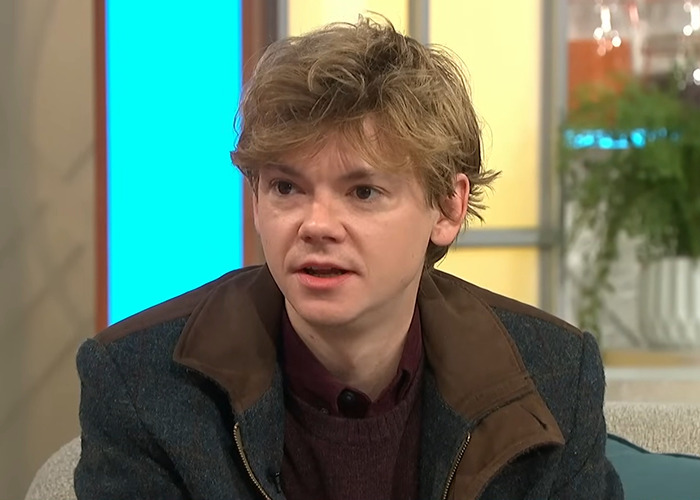 Young man with tousled hair wearing a dark coat during a daytime TV interview discussing viral photos of Sean Connery.
