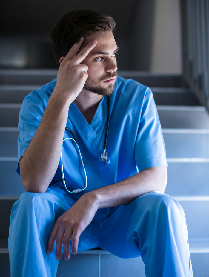 Young male doctor in blue scrubs sitting on stairs, deep in thought about shocking discoveries made during autopsies.