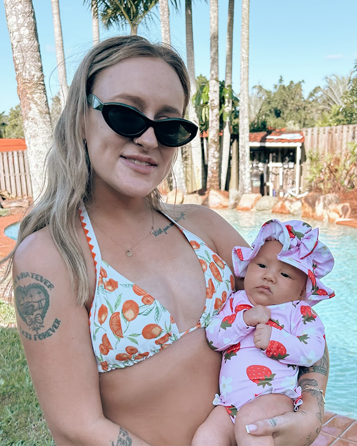 Woman wearing sunglasses holding 5-month-old baby in strawberry swimwear by a pool with palm trees in the background.