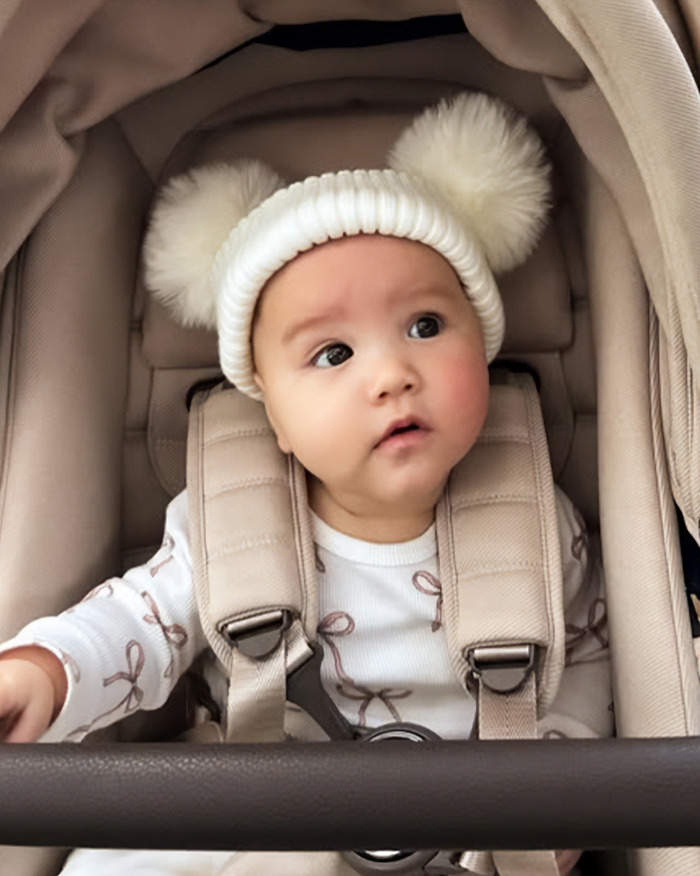 5-month-old baby sitting in a stroller wearing a white hat with pom-poms, strapped in securely and looking to the side