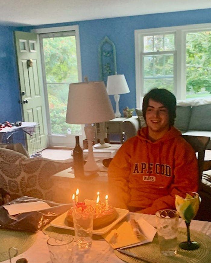 College student sitting at home with birthday cake, candles lit, and late night snack items on the table in cozy living room.