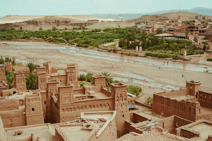 Ancient historical site near a river with adobe buildings and green vegetation in an arid landscape.