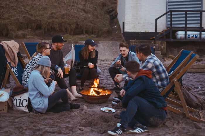 Group of friends sitting around a fire on the beach, sharing laughs and drinks, capturing weird things couples started doing.