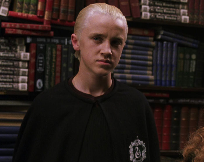 Tom Felton as Draco Malfoy in Slytherin robes standing in front of bookshelves in a Harry Potter scene.