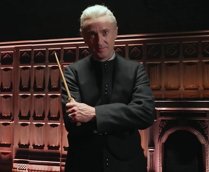 Tom Felton dressed as Draco Malfoy holding a wand on a Harry Potter set with an intense expression.