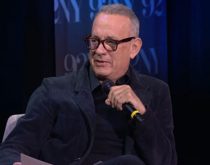 Tom Hanks speaking into a microphone during an interview, addressing childhood confusion violence and deprivation.