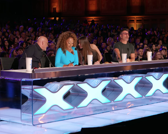 America's Got Talent judges seated behind a panel with illuminated Xs during a live talent show audition.