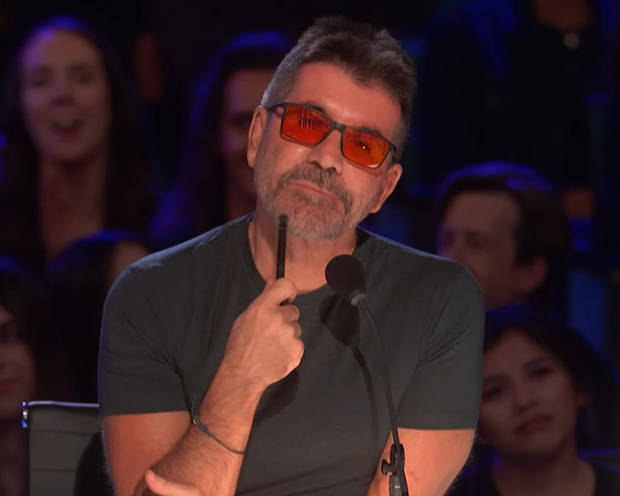 Man wearing orange sunglasses and a black shirt pondering during Americas Got Talent audition panel.
