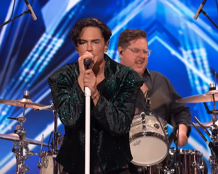 Tom Sandoval performing on stage during Americas Got Talent audition with drummer in the background.