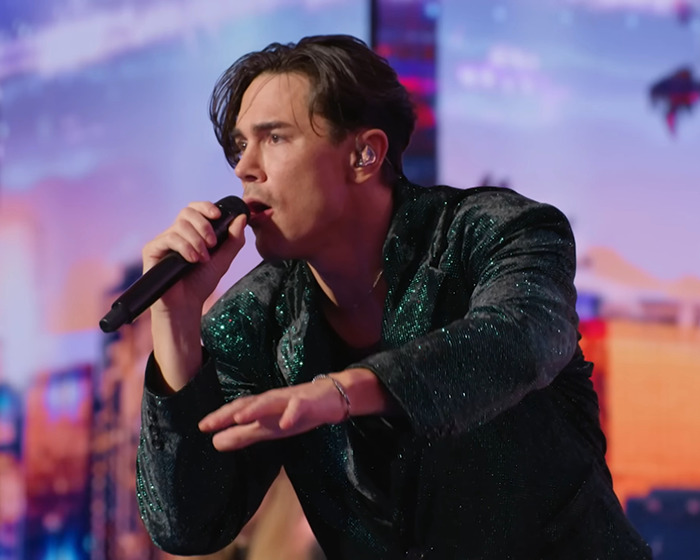 Tom Sandoval performing on Americas Got Talent stage, wearing a sparkly dark jacket and holding a microphone.