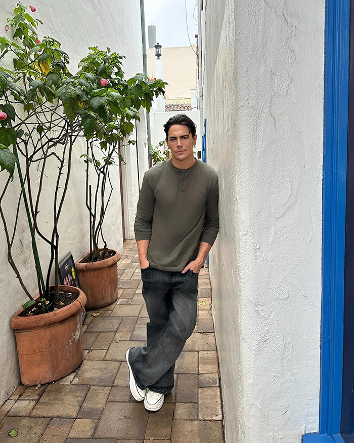 Tom Sandoval leaning against a white wall in a narrow alley with potted plants, related to Americas Got Talent audition.