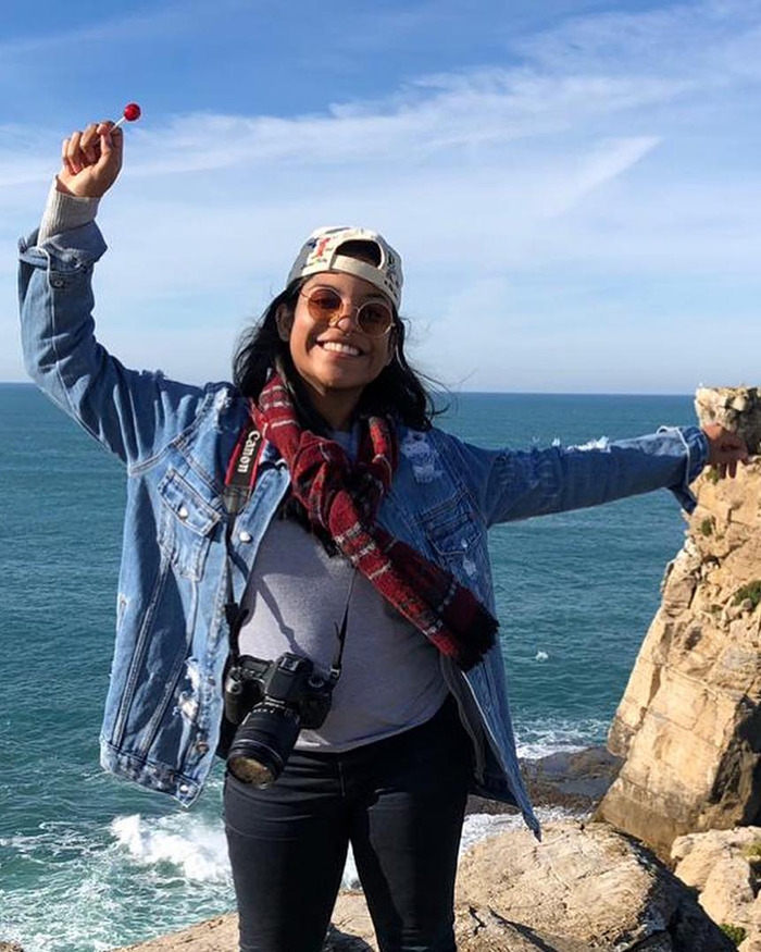 Smiling tourist wearing sunglasses and denim jacket, standing on rocky cliff by ocean, with camera around neck and candy in hand.