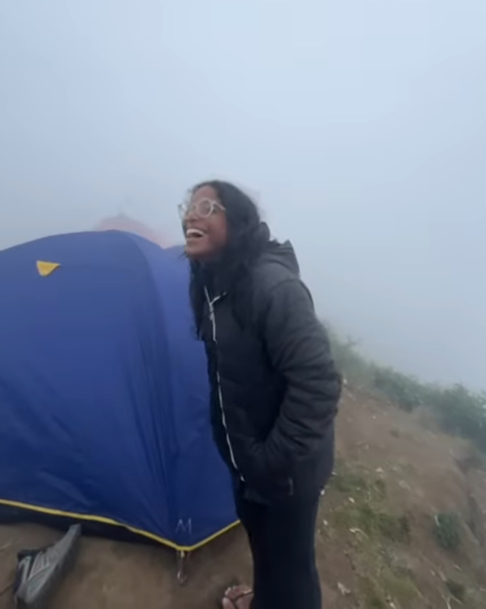 Young tourist smiling near a blue tent in foggy conditions on an active volcano, capturing a final viral moment.