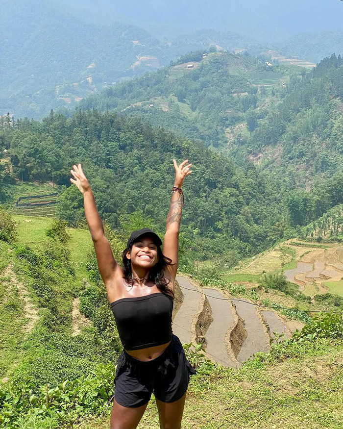 Smiling woman in black outfit raising hands with terraced fields and green mountains in background on a sunny day.